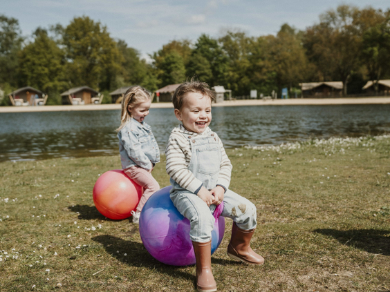 Twee lachende kinderen zitten op een skippybal en stuiteren over het grasveld bij de zwemvijver op Vakantiepark Dierenbos.