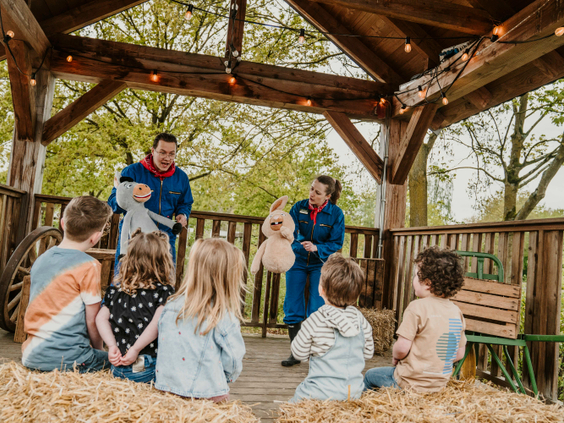 Een groep kinderen zit op strobalen te kijken naar een poppenkastvoorstelling door het animatieteam van Vakantiepark Dierenbos.