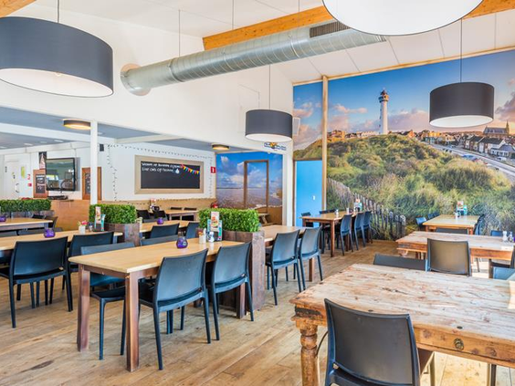 Het sfeervolle restaurant en eetcafé op Roompot Vakantiepark Egmond aan Zee, met houten tafels en een grote wandfoto van de duinen en de vuurtoren.