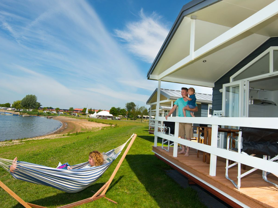 Relaxen in een hangmat bij een chalet aan het water op Vakantiepark Eiland van Maurik, terwijl een gezin op de veranda staat.