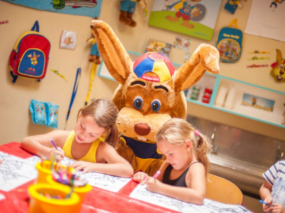 Mascotte Koos Konijn zit gezellig aan tafel met twee meisjes die aan het knutselen zijn in de Kids Club van Ferienresort Cochem.