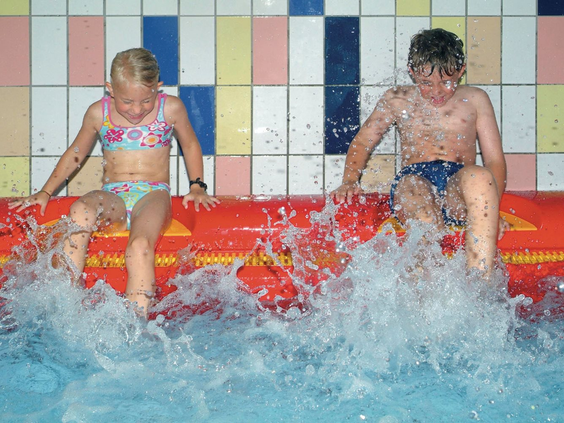 Twee kinderen, een jongen en een meisje, zitten op een rood vlot in het zwembad en spetteren met hun voeten in het water.