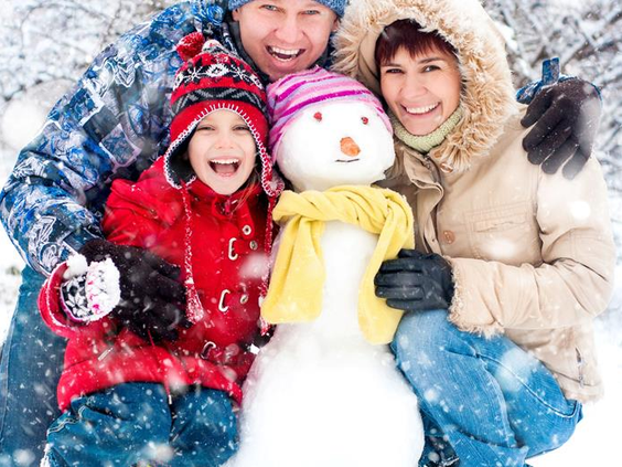 Een lachende familie met kinderen die in de winter een vrolijke sneeuwpop met een gele sjaal knuffelen op Vakantiepark Hasseröder.
