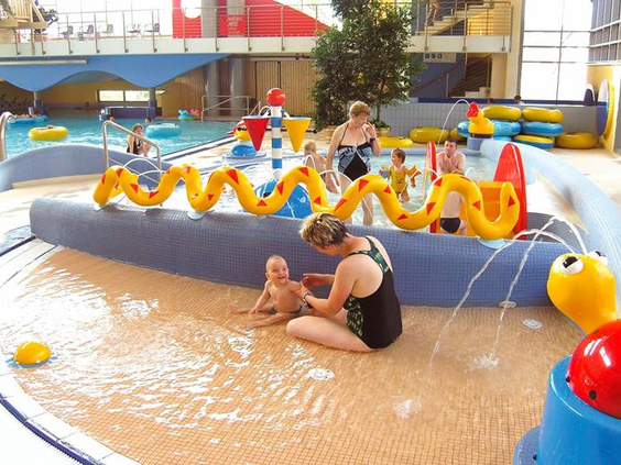 Peuters en kleuters spelen en spetteren vrolijk in het ondiepe kinderbad met een waterslang en andere speeltoestellen.