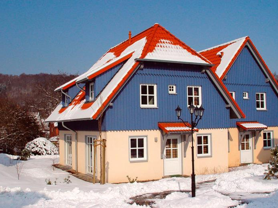 De buitenkant van een gezellig blauw-wit vakantiehuis met een rood, besneeuwd dak op Vakantiepark Hasseröder in de winter.