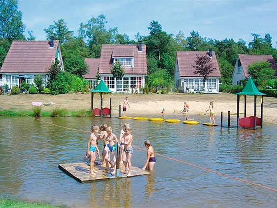 Kinderen spelen op een houten vlot in de zwemvijver van Vakantiepark Hellendoorn in Overijssel, met vakantiehuisjes op de achtergrond.