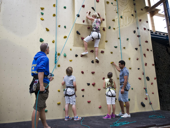 Kinderen en volwassenen bij de indoor klimwand op Vakantiepark Hunzedal, waar een meisje onder begeleiding naar boven klimt.