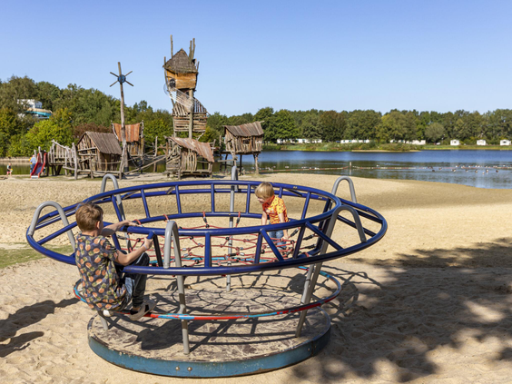 Kinderen spelen in een draaimolen in de zandspeeltuin aan de rand van de recreatieplas op Vakantiepark Hunzedal.