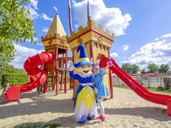 Een vrolijke mascotte poseert bij een houten speelkasteel op de camping