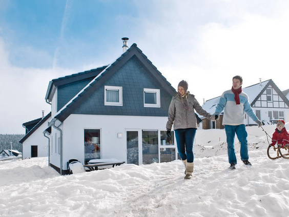 Een familie wandelt hand in hand door de sneeuw naar hun vrijstaande chalet op vakantiepark Landal Winterberg, met een kind op een slee.