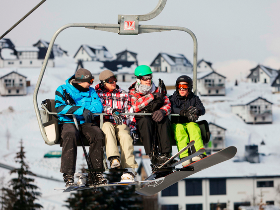 Een gezin met ski's en snowboards zit in de stoeltjeslift boven het vakantiepark Landal Winterberg, met uitzicht op de chalets.