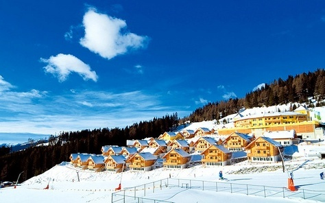 Een prachtig uitzicht op een vakantiepark met houten chalets direct aan de besneeuwde skipiste onder een helderblauwe hemel.