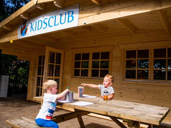 Twee jonge kinderen zitten gezellig te knutselen aan een picknicktafel voor het houten clubhuis van de Kidsclub op Vakantiepark Leuvert.