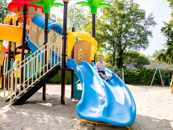 Een jongen glijdt met een lach van de blauwe glijbaan in de zandspeeltuin van Vakantiepark Leuvert.