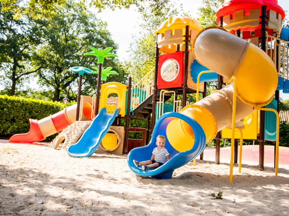 Een kindje glijdt van een kleurrijk speeltoestel in de buitenspeeltuin van Vakantiepark Leuvert, omringd door bomen.