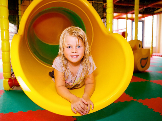 Een lachend meisje ligt onderaan een gele tunnelglijbaan in de binnenspeeltuin van Vakantiepark Marina Beach.