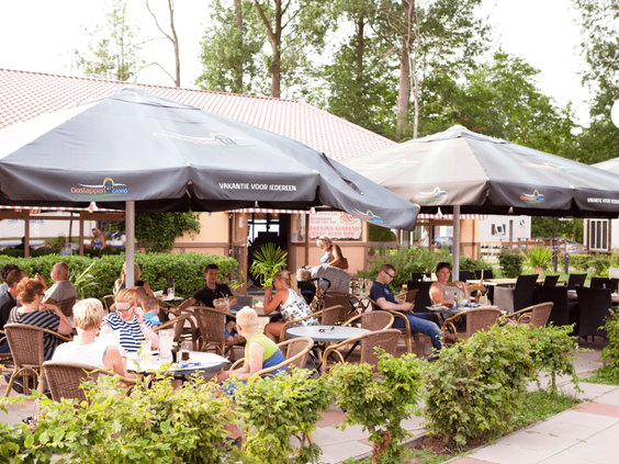 Gezellig druk terras van het restaurant op Vakantiepark Marina Beach, met grote parasols en uitzicht op het groen.