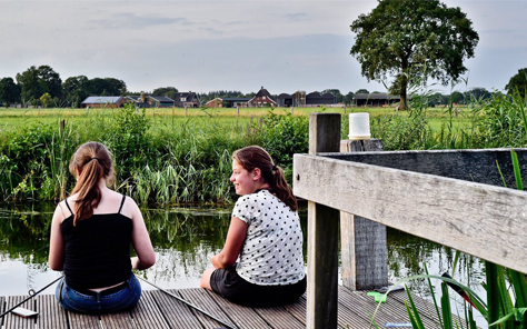 Twee meisjes zitten aan de waterkant op een houten vlonder te genieten van het uitzicht over de rivier bij Vakantiepark Molke.