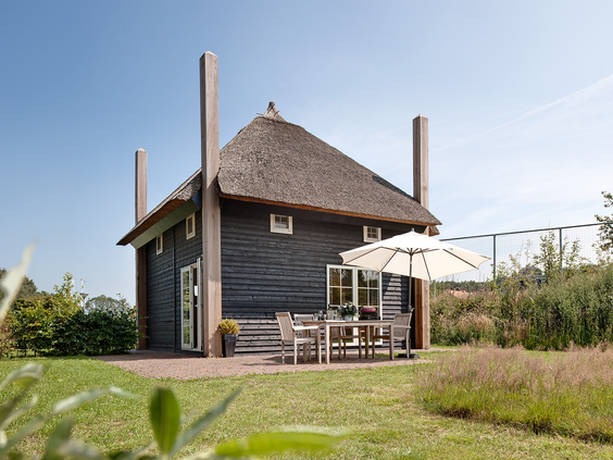 Een luxe, zwarte Regge Lodge met een rieten dak en een zonnig terras met tuinmeubilair op Vakantiepark Molke in Overijssel.