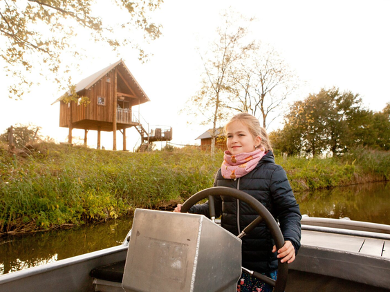 Een meisje staat aan het roer van een fluisterbootje en vaart over de rivier de Regge bij Vakantiepark Molke, met een boomhut op de achtergrond.