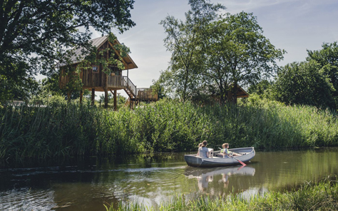Een gezin vaart in een sloep over de rivier de Regge langs de boomhutten van Vakantiepark Molke, omgeven door groene natuur