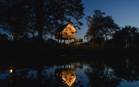 Een sfeervol verlichte boomhut aan het water bij Vakantiepark Molke, weerspiegelend in de rivier de Regge tijdens de schemering.