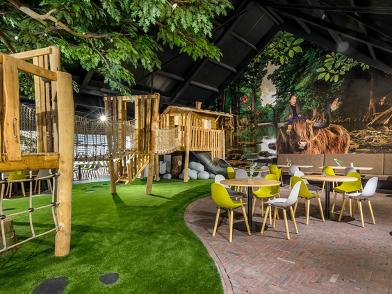 Het indoor speelparadijs van Vakantiepark Schaijk, met houten klimtoestellen en een gezellig terras voor ouders.