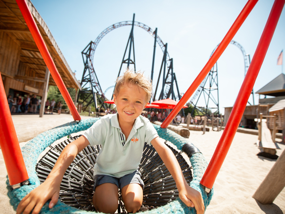 Een jongen speelt op een grote nestschommel in de speeltuin van Vakantiepark Slagharen, met de Gold Rush achtbaan op de achtergrond.