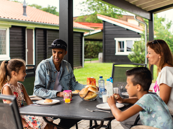 Een gezin geniet samen van een maaltijd op het overdekte terras van hun vakantiehuisje bij Vakantiepark Slagharen.
