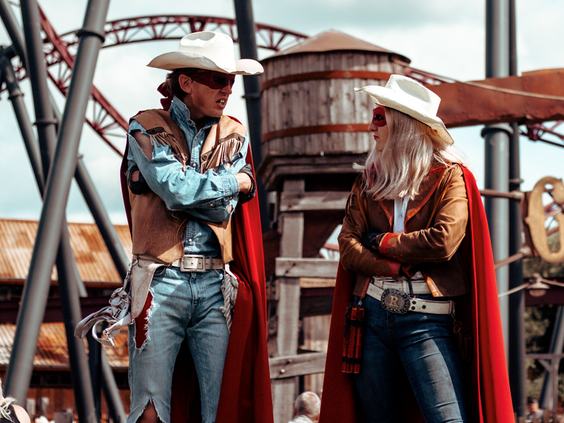 Twee acteurs in cowboykleding met rode capes voeren een stuntshow op met op de achtergrond de Gold Rush achtbaan.