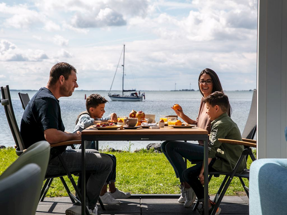 Een gezin met twee kinderen geniet van een maaltijd op het terras van hun vakantiehuis, met uitzicht op het water en een zeilboot.