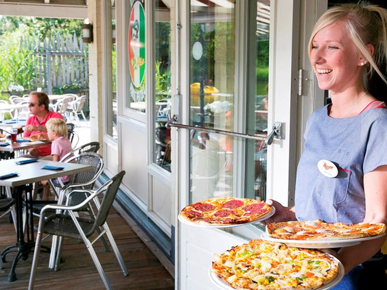 Een medewerker serveert vrolijk versgebakken pizza's bij de pizzeria van Vakantiepark Weerterbergen.