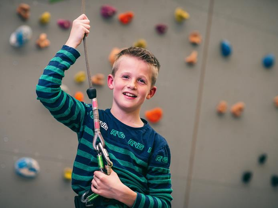 Lachende jongen is gezekerd en klaar om te klimmen op de indoor klimwand vol gekleurde grepen op Vakantiepark Weerterbergen.