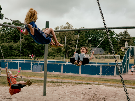 Drie kinderen schommelen samen in de buitenspeeltuin van Vakantiepark Westerbergen met een sportveld op de achtergrond.