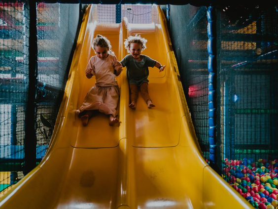 Twee kinderen glijden lachend van de brede gele glijbaan zo de kleurrijke ballenbak in bij de indoor speeltuin van Westerbergen.
