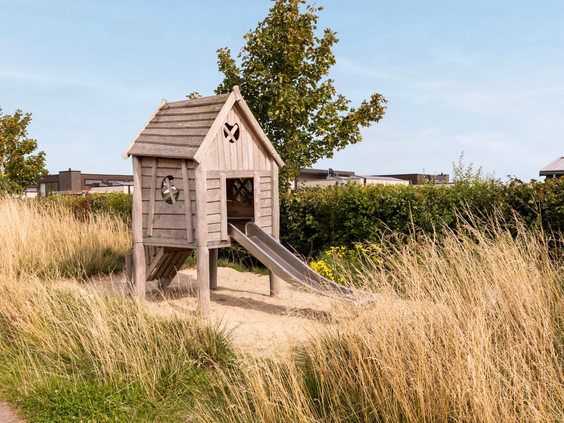 Houten speeltoestel met een glijbaan in een natuurspeeltuin met hoog gras op Vakantiepark Zeedijk in Zuid-Holland.