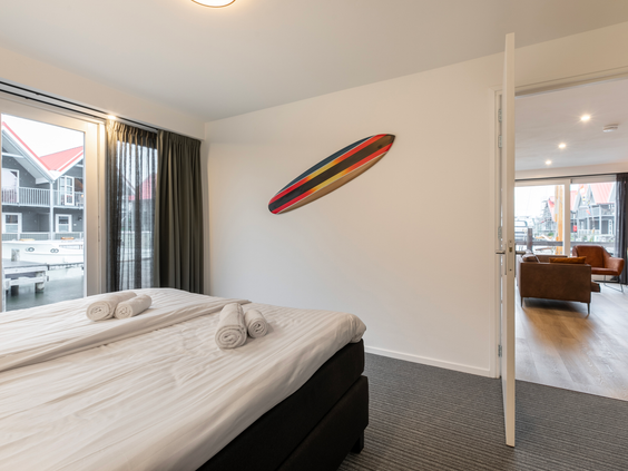 Sfeervolle slaapkamer met een opgemaakt bed en een decoratieve surfplank aan de muur, met uitzicht op het water.