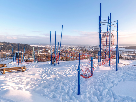 Een buitenspeeltuin met blauwe klimrekken bedekt met sneeuw, met een prachtig uitzicht over het winterse landschap van Winterberg.