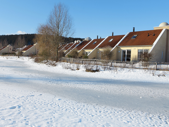 Een rij cottages van Center Parcs Park Hochsauerland aan een bevroren, met sneeuw bedekt meer op een zonnige winterdag.