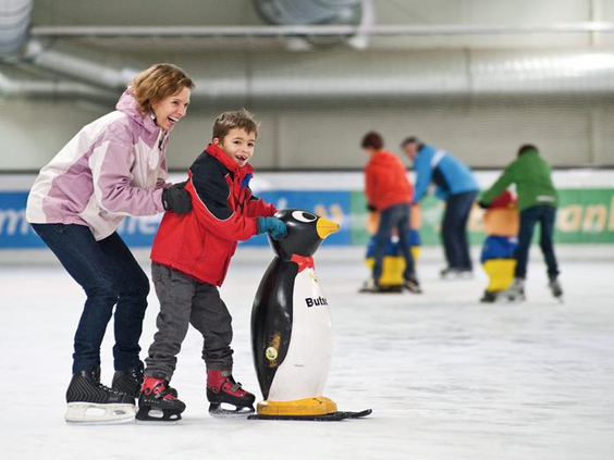 Een kind leert schaatsen met een pinguïn-schaatshulp op de overdekte ijsbaan van Landal Salztal Paradies, geholpen door een ouder.