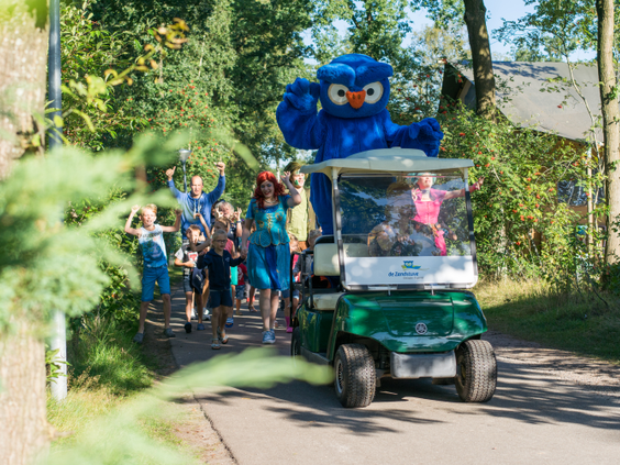 Parade van vrolijke kinderen achter een golfkarretje met daarop de blauwe uilenmascotte cobus van de zandstuve