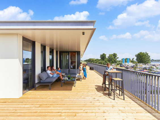 Een gezin ontspant op het zonnige en ruime houten terras van een moderne waterlodge direct aan het water in Zeewolde.