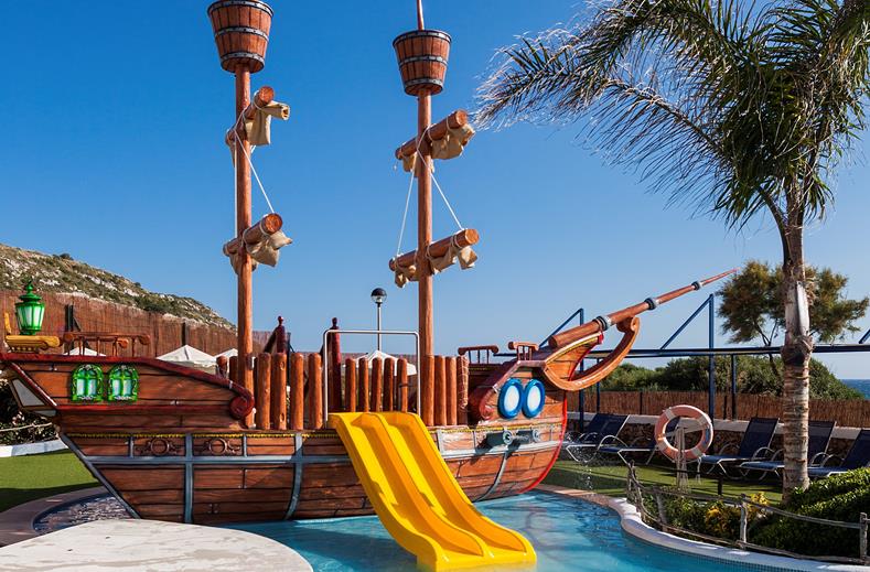 Groot houten piratenschip speeltoestel met een gele glijbaan in het kinderbad van familiehotel Royal Son Bou op Menorca.