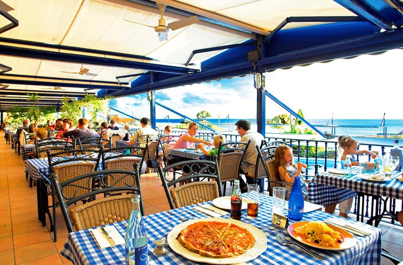 Gasten genieten van een lunch, waaronder pizza, op het sfeervolle buitenterras van het restaurant bij Royal Son Bou, met prachtig uitzicht over de zee.