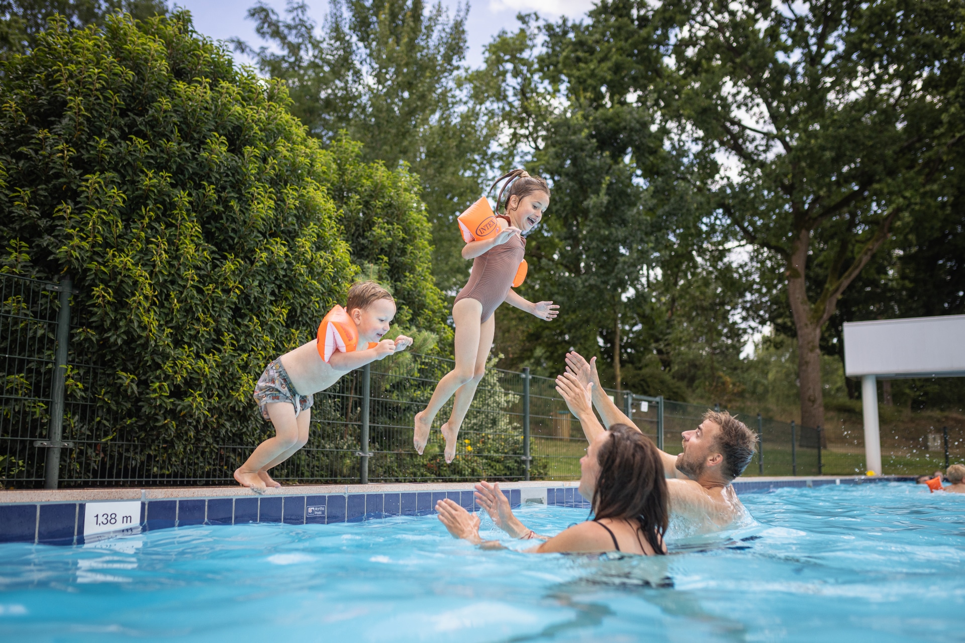 Twee kinderen met zwemvleugels springen in het buitenzwembad, waar hun ouders ze opwachten, op een zonnige dag bij De Katjeskelder.