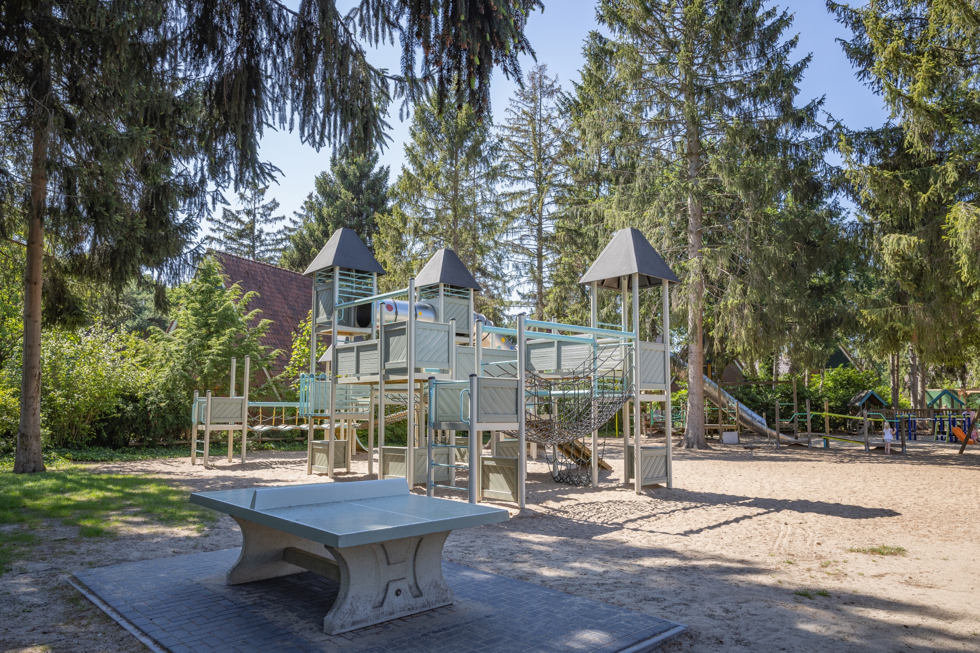 Een groot houten speelkasteel met glijbanen en een tafeltennistafel in de avontuurlijke buitenspeeltuin van De Katjeskelder, omgeven door bomen.