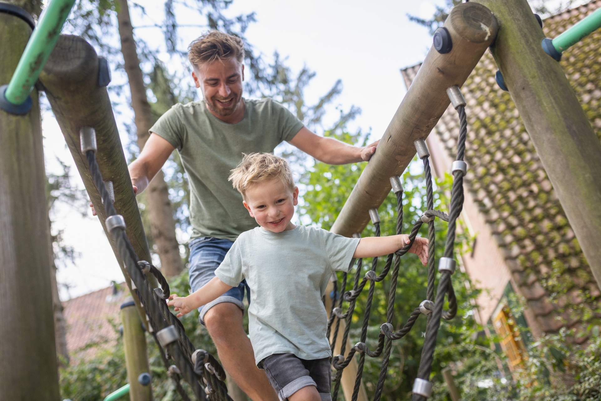 Een vader helpt zijn zoontje balanceren op een wiebelende touwbrug in de avontuurlijke speeltuin van Bungalowpark De Katjeskelder.