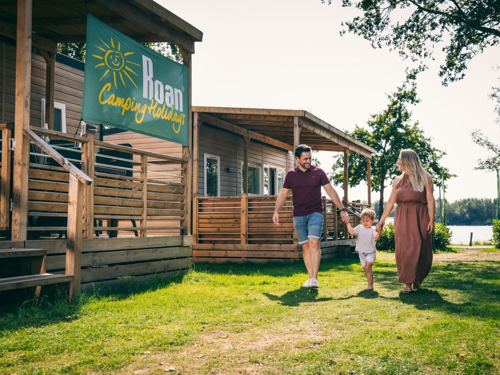 Gezin wandelt hand in hand voor hun houten stacaravan op een grasveld van Camping de Schatberg.
