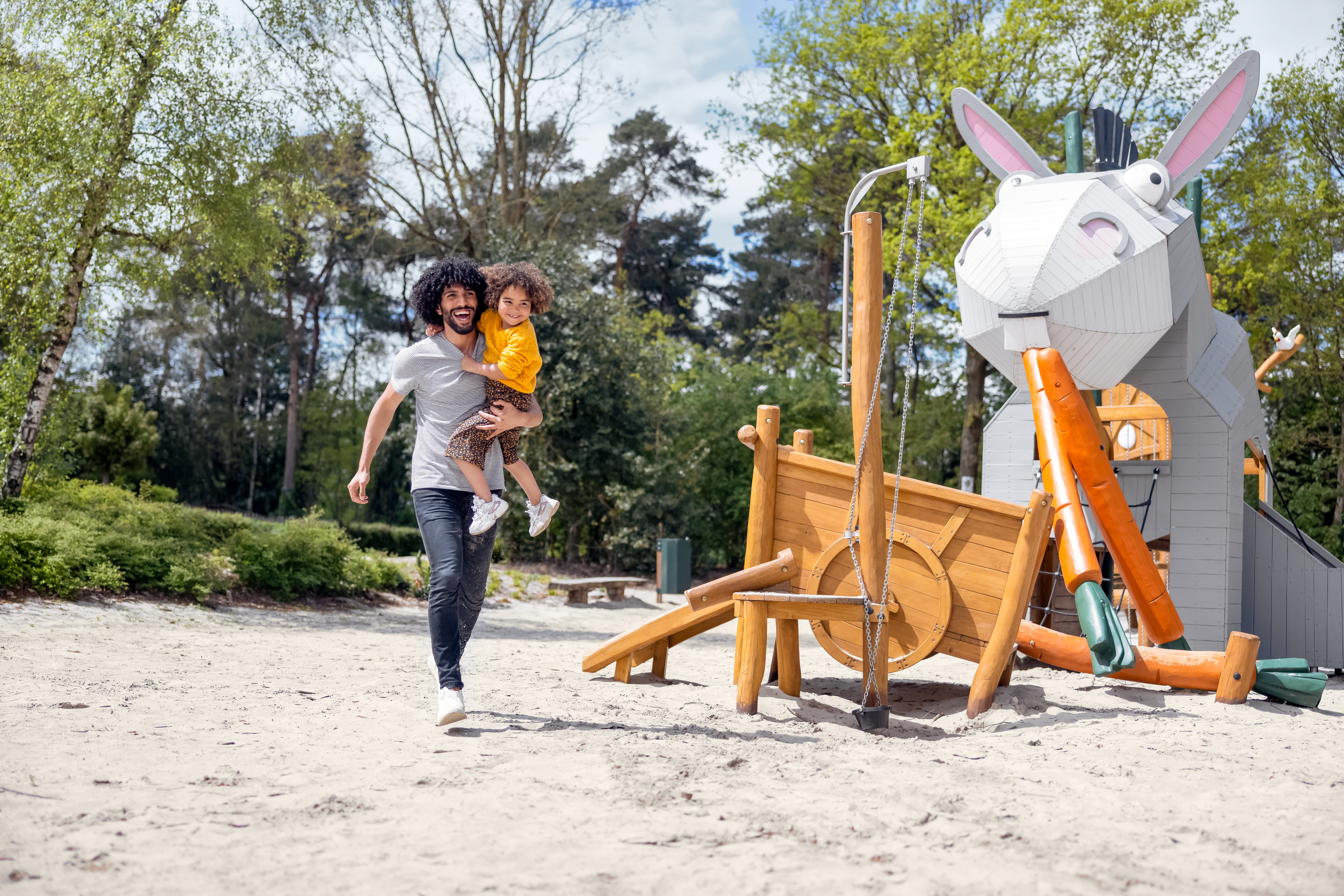 Een vader rent met zijn kind in de armen door een zandspeeltuin bij Center Parcs Erperheide, met een groot speeltoestel in de vorm van een konijn.