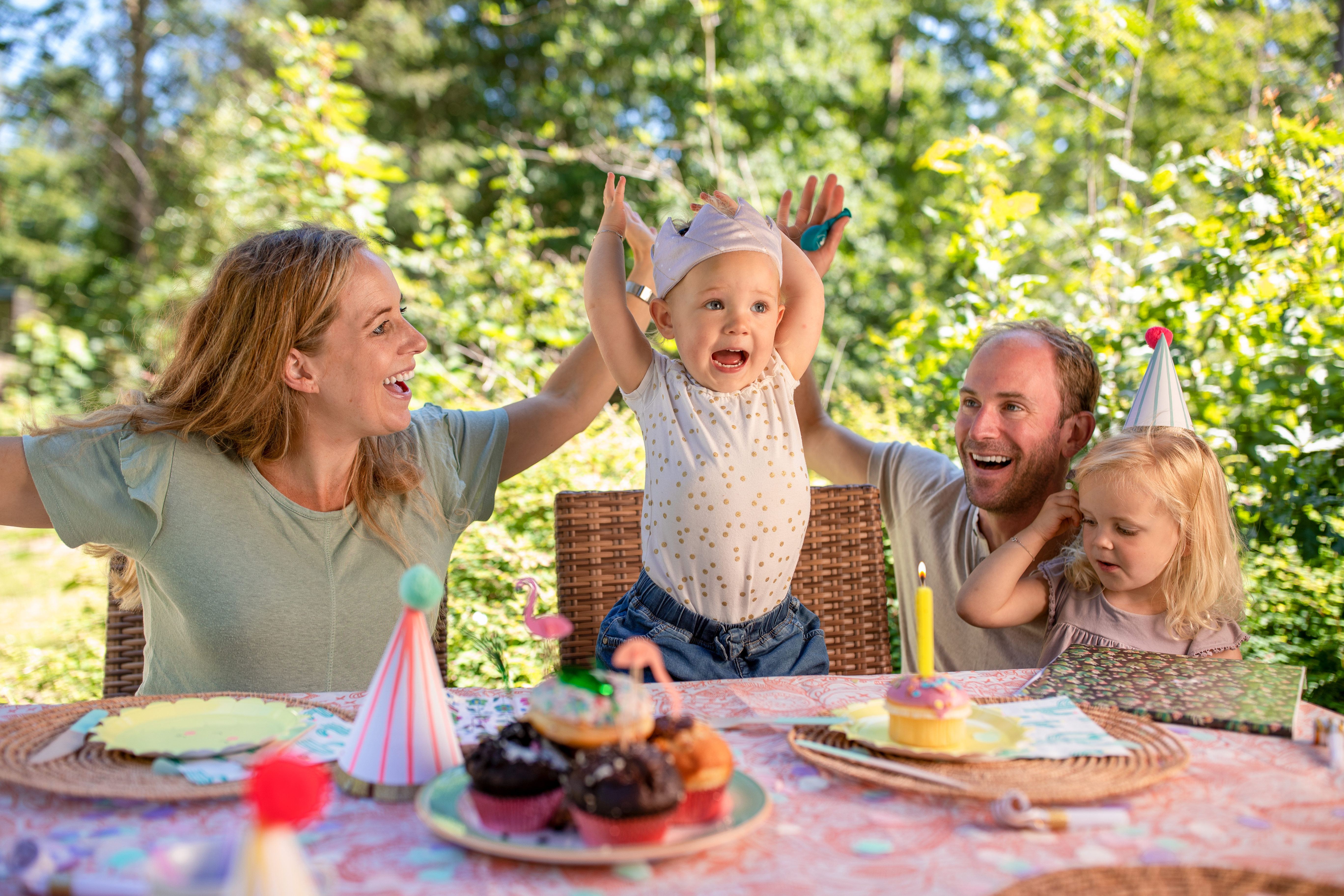 Een gezin met twee jonge kinderen viert een verjaardag aan een versierde picknicktafel in het bos, met cupcakes en feestmutsen, bij Center Parcs Erperheide.